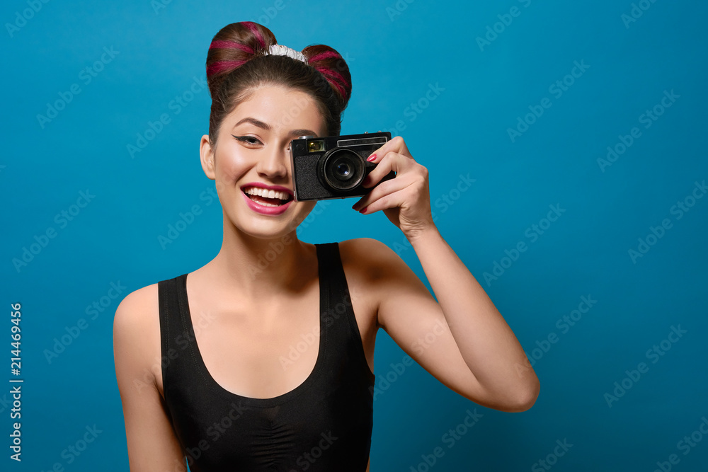 Smiling, happy girl taking photo with old camera. Laughing, keeping camera, having fancy hairstyle with a bow, colorful day make up with pink lipstick. Posing on saturated deep blue background. Smiling, happy girl taking photo with old camera. Laughing, keeping camera, having fancy hairstyle with a bow, colorful day make up with pink lipstick. Posing on saturated deep blue background.
