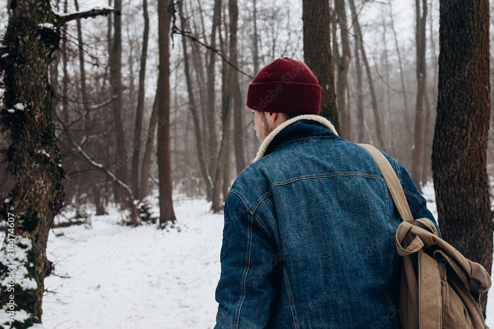 stylish hipster traveler with backpack in red hat walking in winter snowy forest. wanderlust and adventure concept with space for text. back view. atmospheric moment