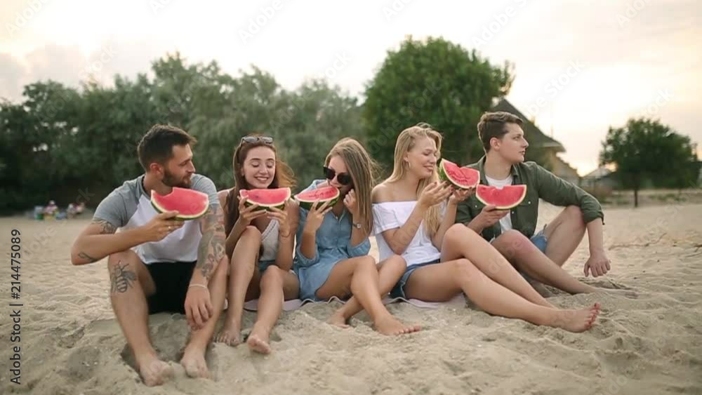 Happy Friends Eating Watermelon Sitting on Sandy Beach on Vacation. Young Men and Women Wearing Blue Jeans Shorts. Friendship and Summer Concept Happy Friends Eating Watermelon Sitting on Sandy Beach on Vacation. Young Men and Women Wearing Blue Jeans Shorts. Friendship and Summer Concept
