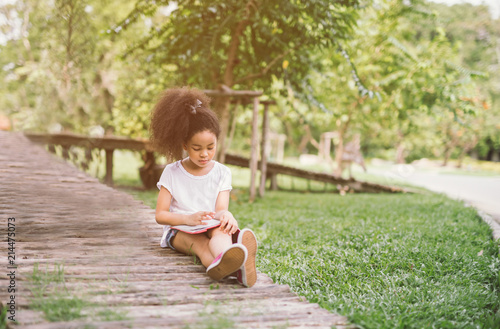 little Afro child girl reading book between green spikes meadow garden.