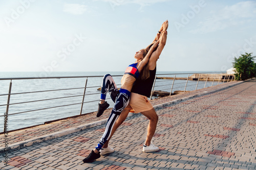 Young couple doing sports exercises for body together, wearing sportswear, while working out near the sea, on the quay. Healthy lifestyle.