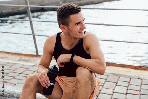 Young sportsman holding a bottle of water while sitting on the ground, after workout outdoors, near the sea. Healthy lifestyle.