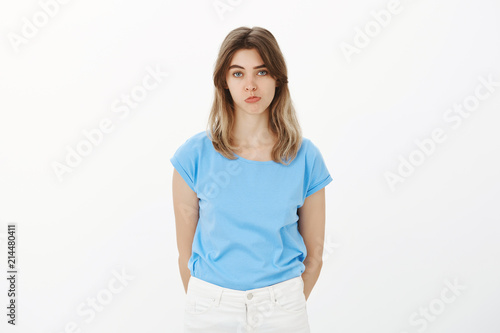 Portrait of unhappy cute female coworker in blue t-shirt, pouting and sulking, gazing at camera with lack of interest, being indifferent and unimpressed, standing upset over gray background