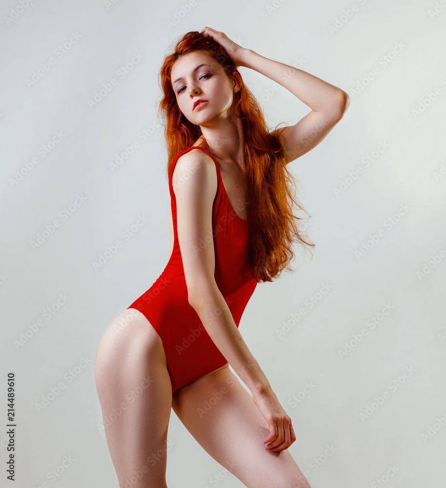 Young sexy slim woman in red swimsuit posing in studio. Full length fashion  portrait of beautiful girl with long wavy red hair. Swimwear or bikini  mode. Copy space, gray background Stock Photo