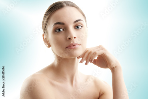 Portrait of a beautiful young woman with naked shoulders smiling while posing at isolated light blue background. Face beauty skin care. Natural make up