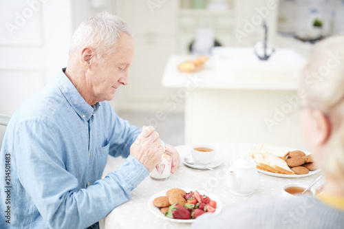 Aged man sitting by table in the kitchen and eating milk dessert with fresh strawberries