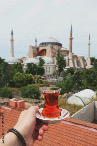 Traditional Turkish tea in a glass on a plate in the hands of a man with a mosque Ayasofya background. 