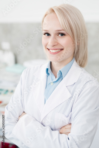 Happy young doctor in whitecoat crossing her arms on ches and looking at you with toothy smile