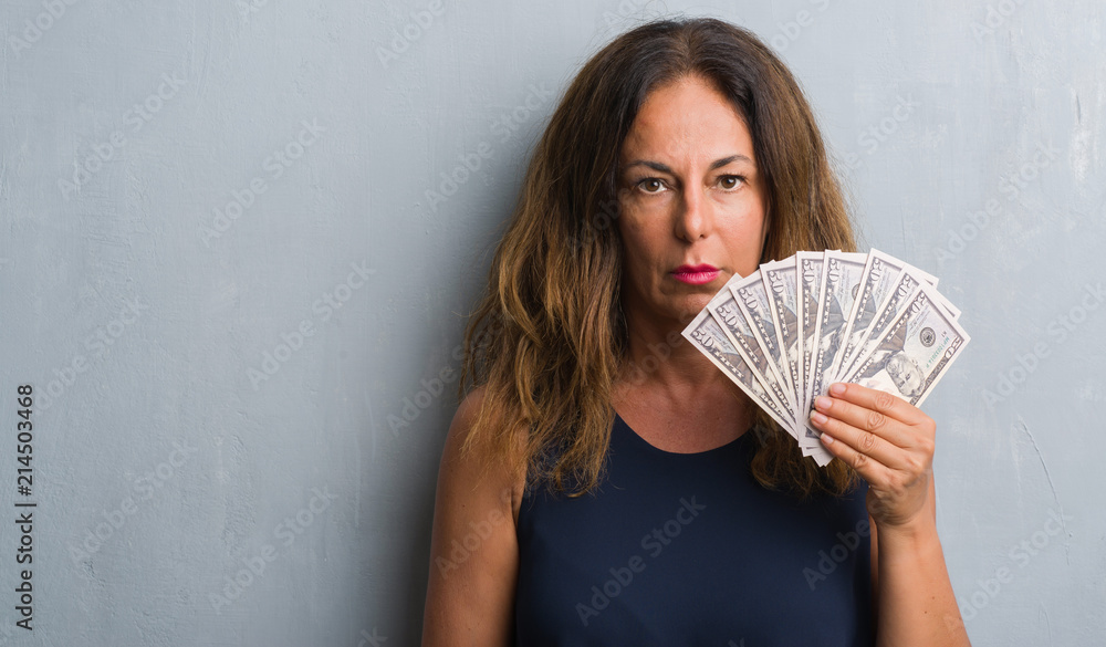 Middle age hispanic woman standing over grey grunge wall holding dollars with a confident expression on smart face thinking serious Middle age hispanic woman standing over grey grunge wall holding dollars with a confident expression on smart face thinking serious