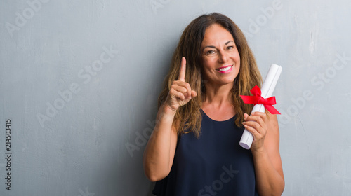 Middle age hispanic woman standing over grey grunge wall holding diploma surprised with an idea or question pointing finger with happy face, number one