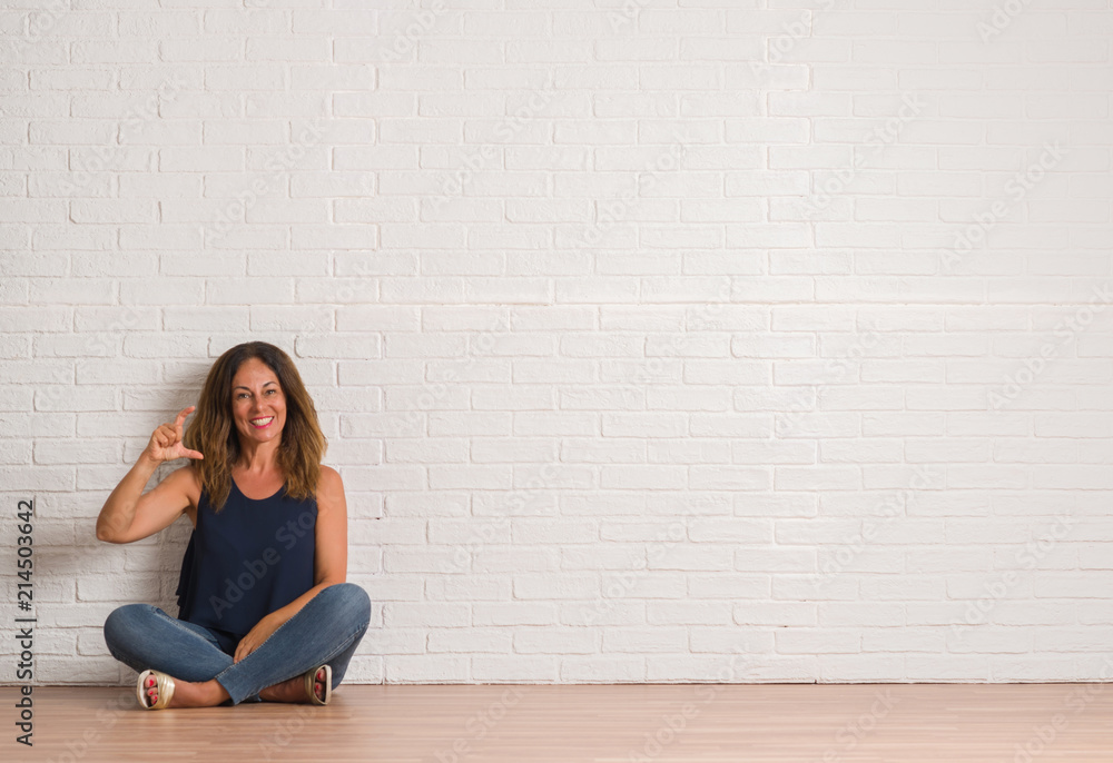 Middle age hispanic woman sitting on the floor over white brick wall smiling and confident gesturing with hand doing size sign with fingers while looking and the camera. Measure concept. Middle age hispanic woman sitting on the floor over white brick wall smiling and confident gesturing with hand doing size sign with fingers while looking and the camera. Measure concept.