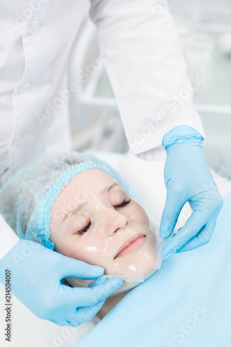 Cosmetician in gloves taking transparent fabric purifying mask off relaxed client face