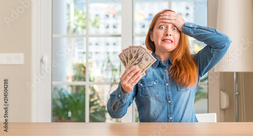 Redhead woman holding dollar bank notes at home stressed with hand on head, shocked with shame and surprise face, angry and frustrated. Fear and upset for mistake.