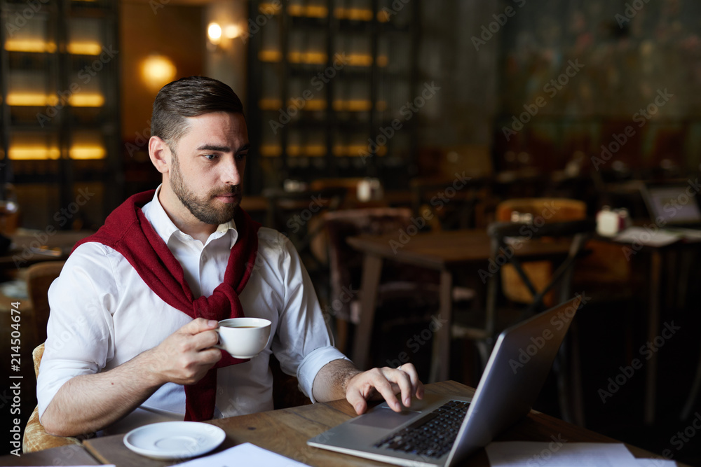 Serious businessman with drink sitting by table in front of laptop and searching for necessary data