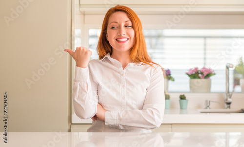 Redhead woman at kitchen smiling with happy face looking and pointing to the side with thumb up.