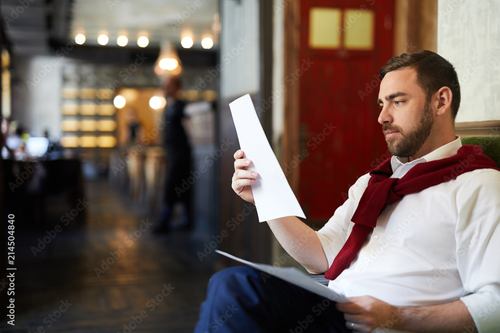 Young elegant man sitting in cafeteria and reading data in document or contract Young elegant man sitting in cafeteria and reading data in document or contract