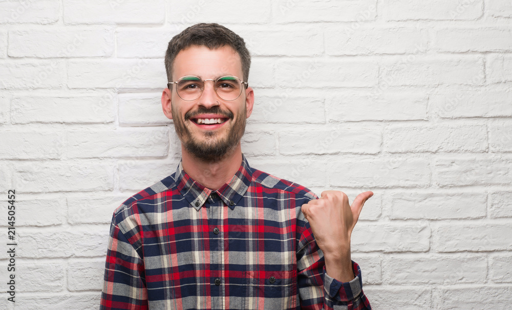 Young adult man standing over white brick wall pointing and showing with thumb up to the side with happy face smiling Young adult man standing over white brick wall pointing and showing with thumb up to the side with happy face smiling