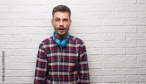 Young adult man over brick wall wearing headphones scared in shock with a surprise face, afraid and excited with fear expression