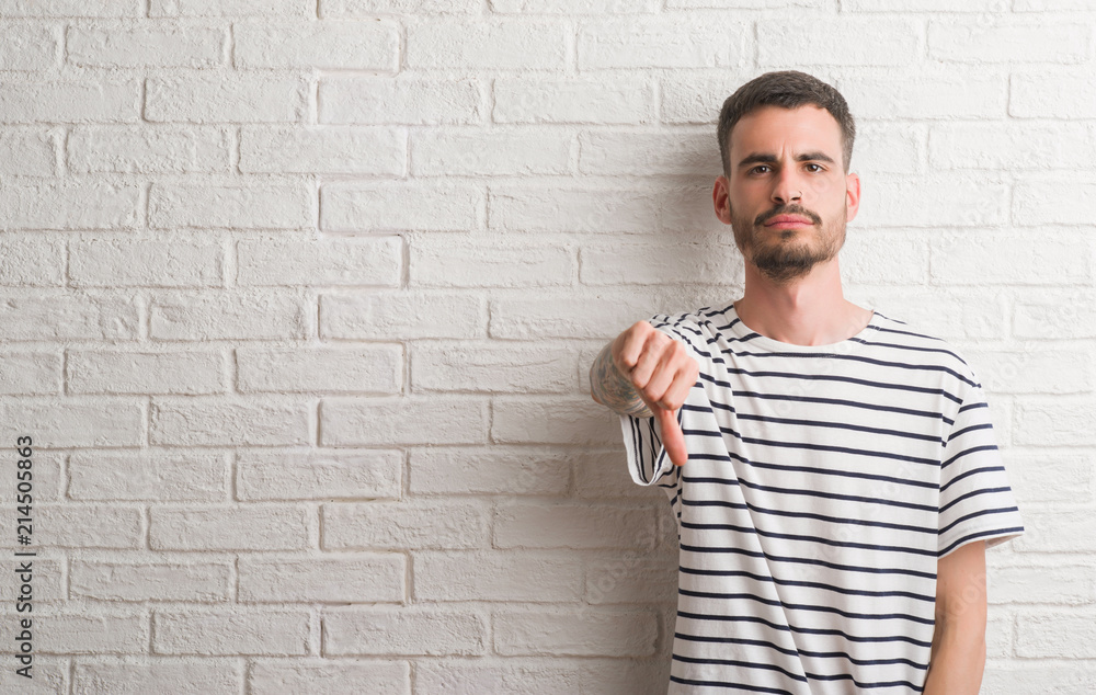 Young adult man standing over white brick wall with angry face, negative sign showing dislike with thumbs down, rejection concept Young adult man standing over white brick wall with angry face, negative sign showing dislike with thumbs down, rejection concept