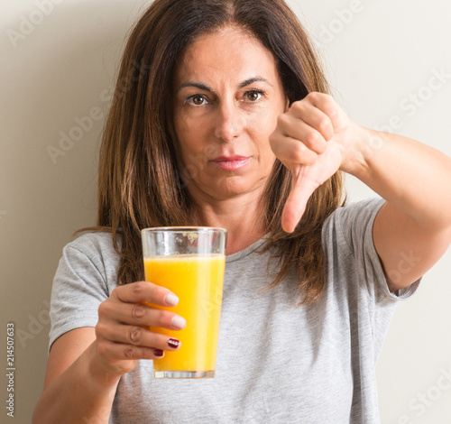 Middle age woman drinking orange juice in a glass with angry face, negative sign showing dislike with thumbs down, rejection concept photo