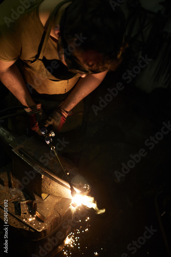 Young blacksmith professional in workwear standing by anvil and welding workpiece