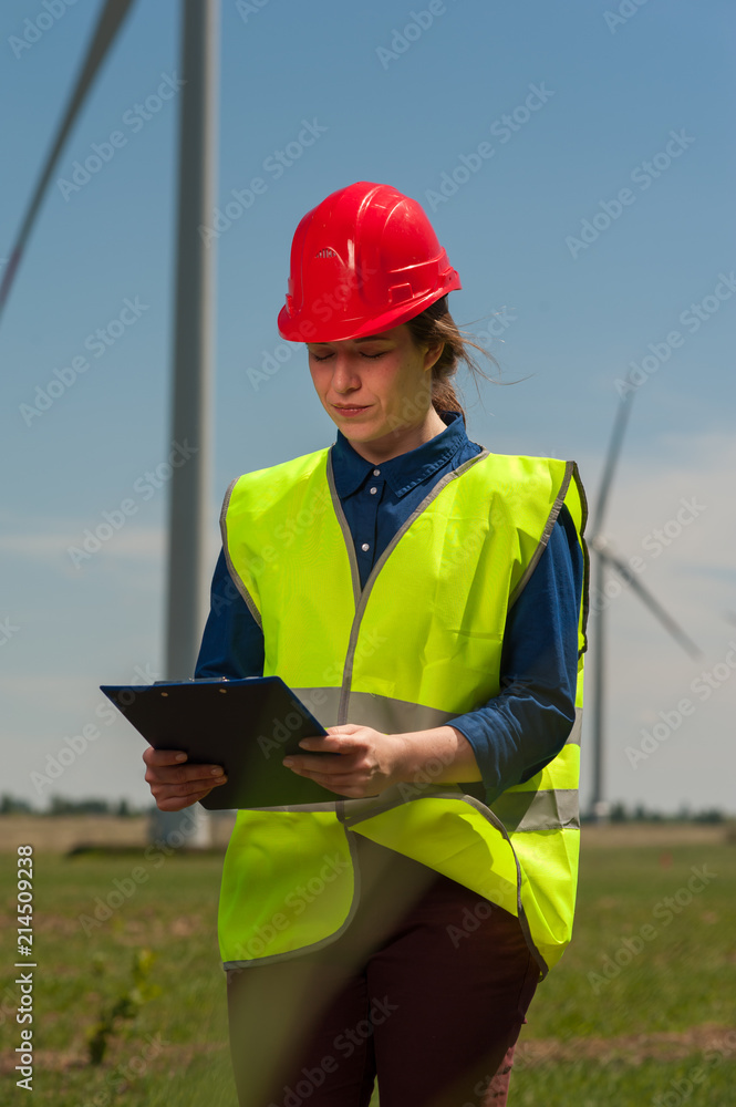 Portrait of a serious successful young female engineer in a green vest and red hard hat designing a plan against a background of windmills Portrait of a serious successful young female engineer in a green vest and red hard hat designing a plan against a background of windmills