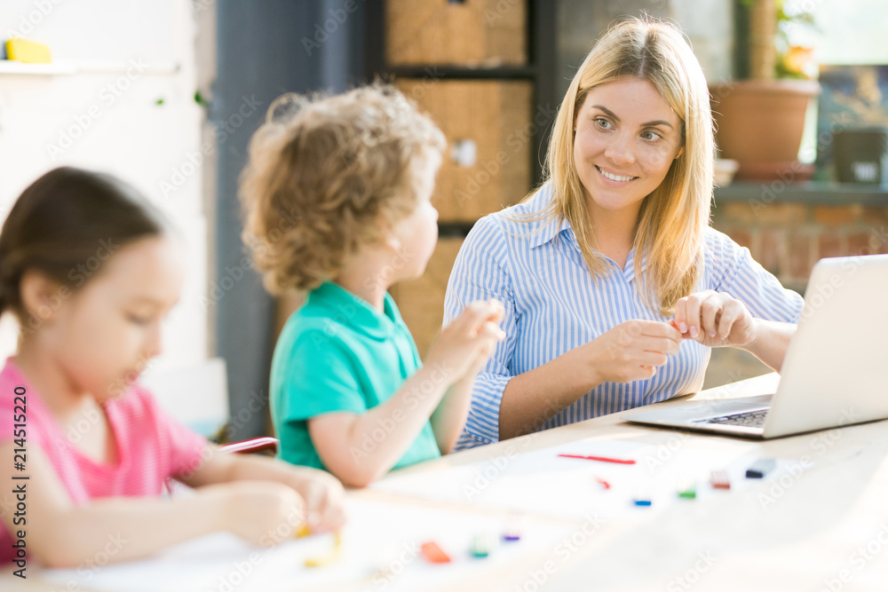 Smiling teacher sitting at the table with laptop and sculpting from play clay together with pupils Smiling teacher sitting at the table with laptop and sculpting from play clay together with pupils