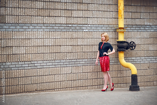 A blonde girl in the fashion style in a red mini skirt and a black blouse walks down the city street in the evening.