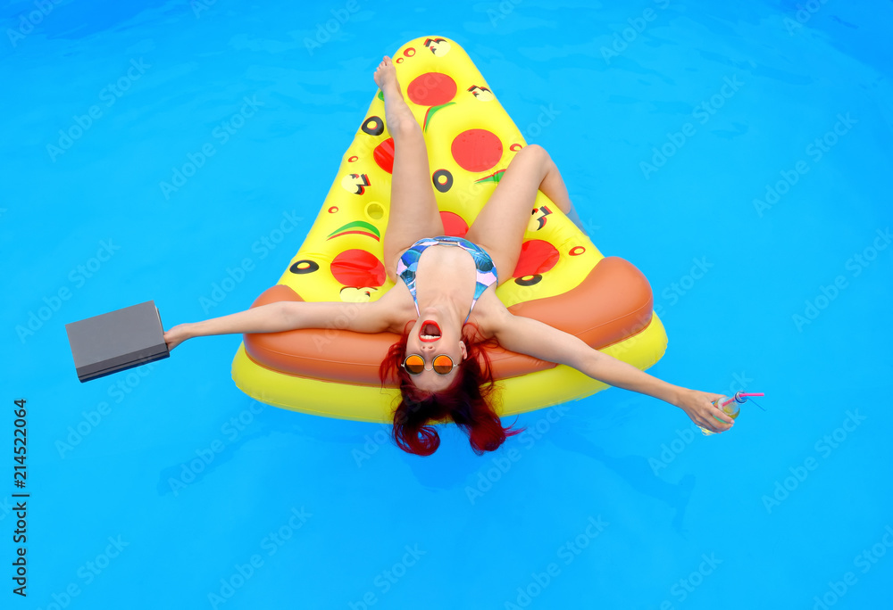 A young woman wearing a bathing suit lies relaxed on an  inflatable swim bed. She takes time out to read a book while she enjoys the pool atmosphere.