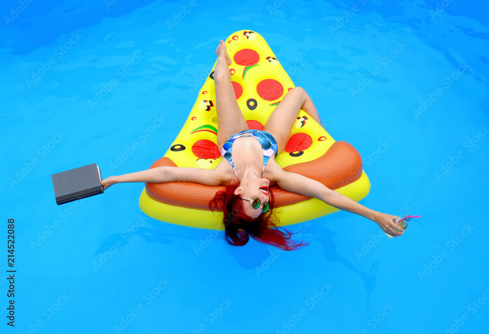 A young woman wearing a bathing suit lies relaxed on an  inflatable swim bed. She takes time out to read a book while she enjoys the pool atmosphere.