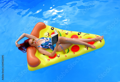 A young woman wearing a bathing suit lies relaxed on an inflatable swim bed. She takes time out to read a book while she enjoys the pool atmosphere.