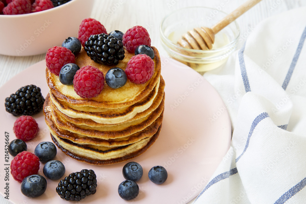 Pancakes with berries and honey on a pink plate over white wooden background, side view. Close-up. Pancakes with berries and honey on a pink plate over white wooden background, side view. Close-up.