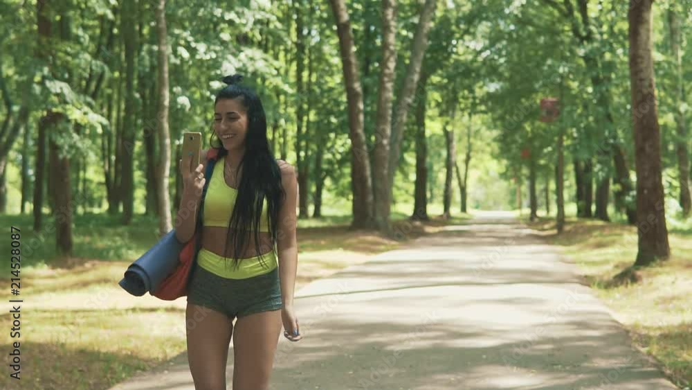 Beautiful brunette fitness woman walking with yoga mat and phone at park. Cute girl in green sportswear chatting before running. Technology concept in slow motion witj copy space Beautiful brunette fitness woman walking with yoga mat and phone at park. Cute girl in green sportswear chatting before running. Technology concept in slow motion witj copy space