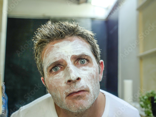 young funny attractive man at home bathroom looking at toilet mirror applying facemask in his face finding himself weird and ugly with the white mask