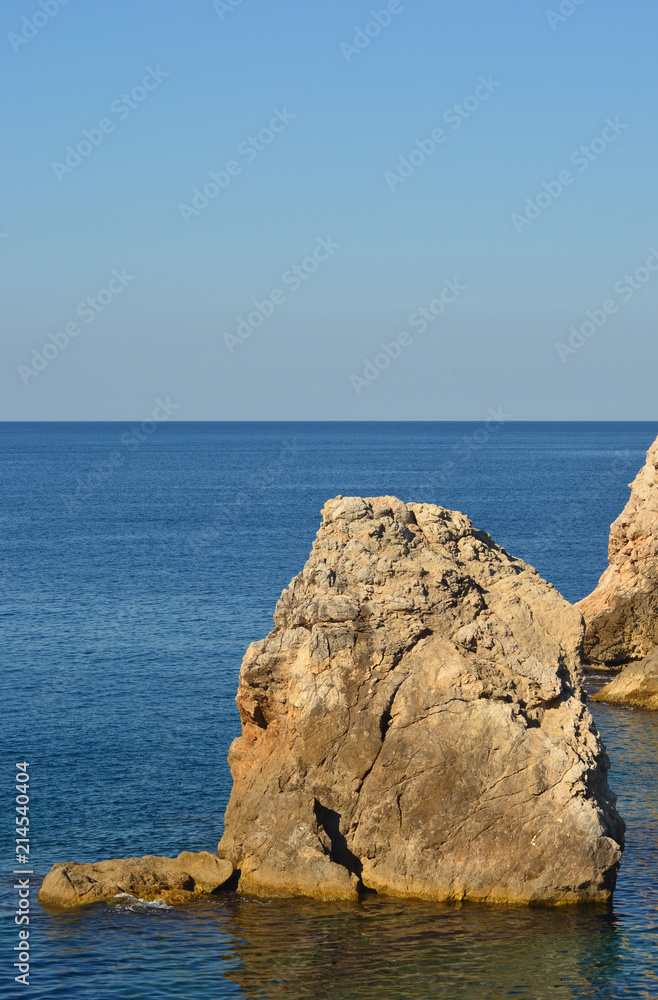 A jagged rock rising out of a blue ocean. Behind is a clear blue sky.