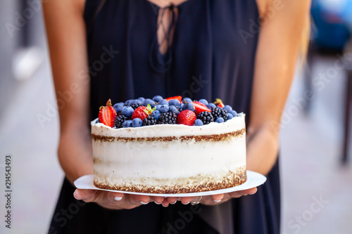 Fototapeta The girl is holding a cake with forest berries, strawberries, blueberries, on the street in the city