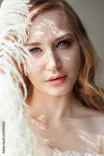 Fototapeta Naklejka Na Ścianę i Meble -  Girl in white light dress and curly hair with big feather near her face, portrait woman with feather, purity and innocence. Curly blonde looks romantic, beautiful eyes. White ostrich feather in hands