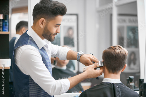 Barber doing new haircut for young client sitting in front of mirror. Wearing white casual shirt, grey waistcoat, watch. Looking concentrated, loving his job. Model covered with special black cape.