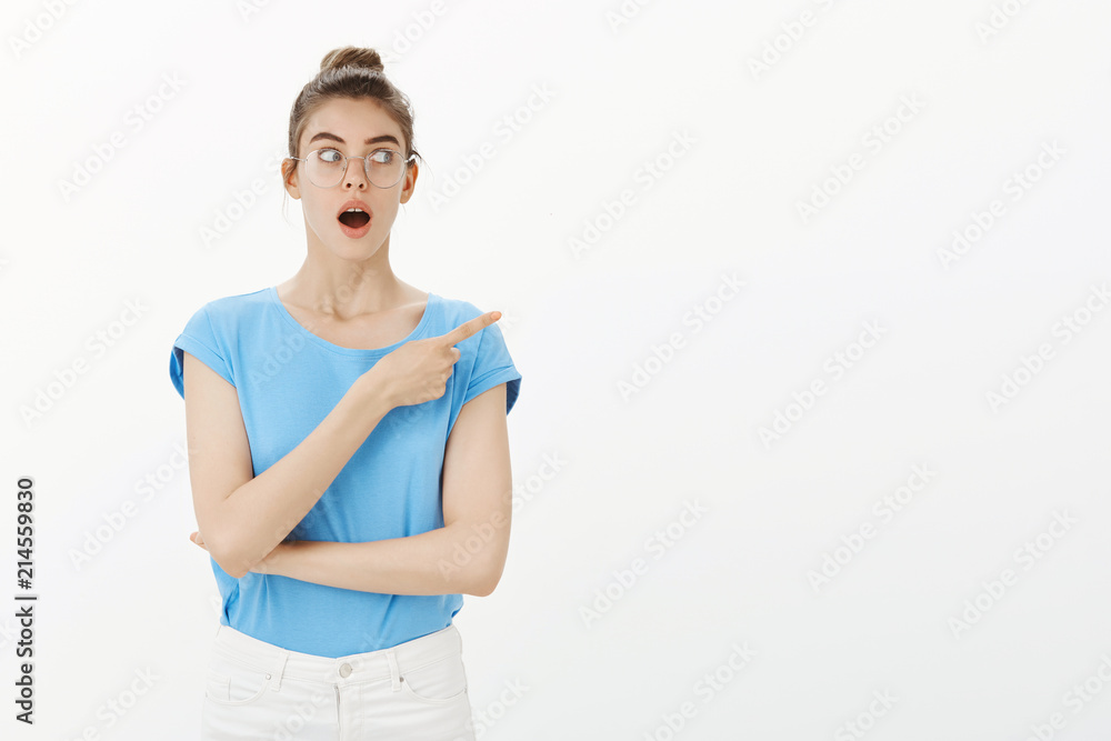 Did you heard new rumor about her. Portrait of shocked and amazed charming woman in glasses and blue t-shirt, pointing right and dropping jaw, gazing with interest while gossiping over grey wall Did you heard new rumor about her. Portrait of shocked and amazed charming woman in glasses and blue t-shirt, pointing right and dropping jaw, gazing with interest while gossiping over grey wall