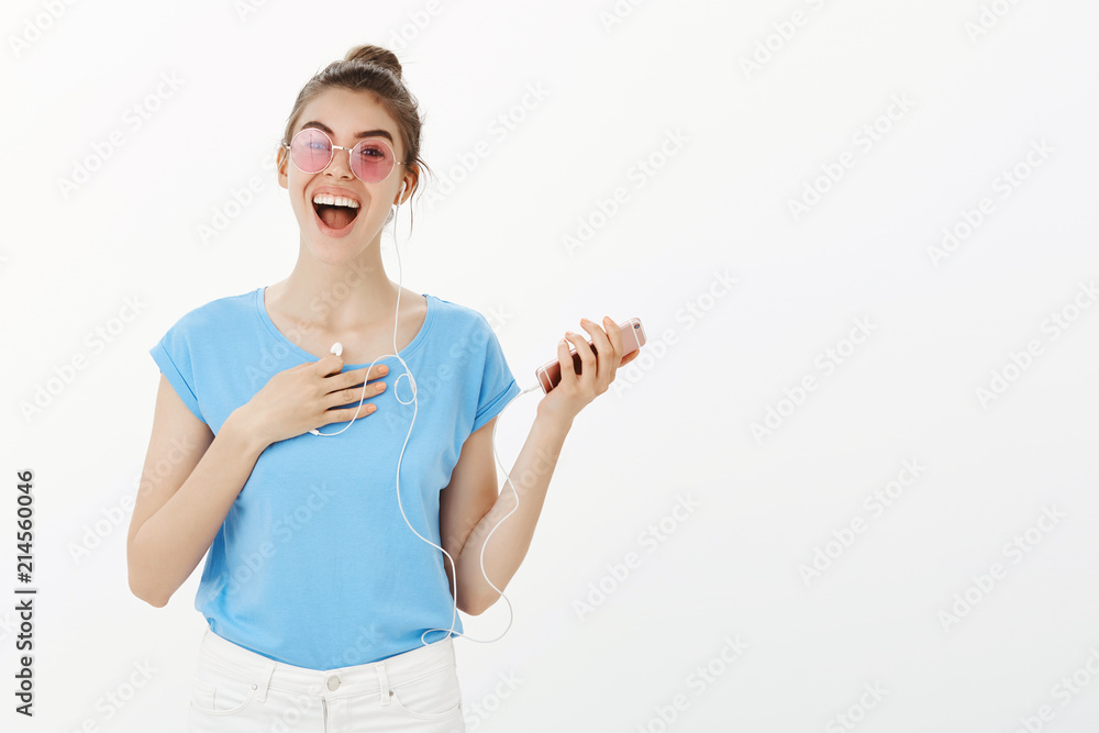 Girl walking on street meeting friend. Portrait of positive surprised attractive woman in pink sunglasses and t-shirt, holding smartphone and earphones, touching chest with arm, smiling broadly Girl walking on street meeting friend. Portrait of positive surprised attractive woman in pink sunglasses and t-shirt, holding smartphone and earphones, touching chest with arm, smiling broadly