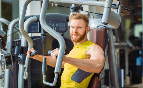 Portrait of a handsome bodybuilder smiling and looking at camera while exercising at a modern fitness machine for pectoral fly and deltoid workout