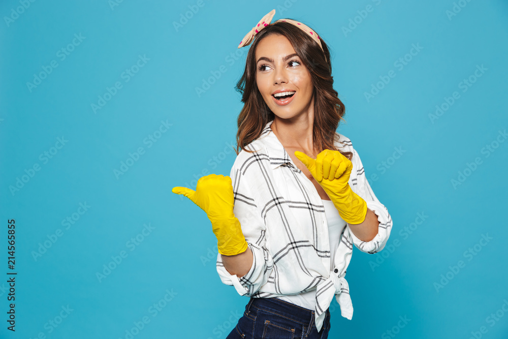 Caucasian housewife 20s pointing fingers aside at copyspace while wearing yellow rubber gloves for hands protection during cleaning, isolated over blue background Caucasian housewife 20s pointing fingers aside at copyspace while wearing yellow rubber gloves for hands protection during cleaning, isolated over blue background