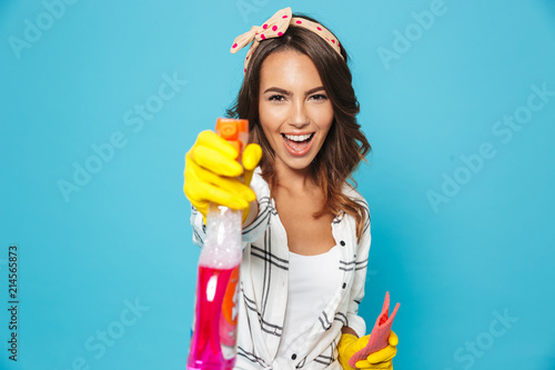 Photo of brunette woman 20s spraying detergent and holding sponge while cleaning in yellow rubber gloves for hands protection, isolated over blue background photo