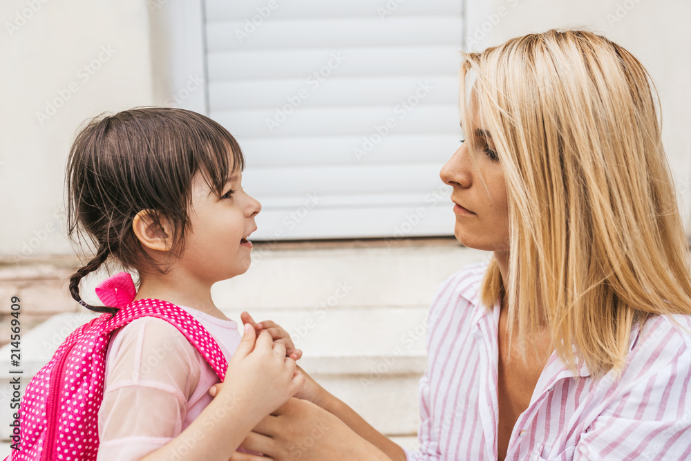 Side view of mother preparing her kid daughter to going to kindergarten sitting on stairs against home outdoor. Portrait of cute little girl pupil with mom before the first preschool day against home