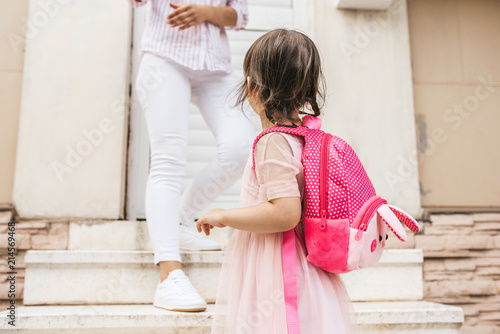 Image of mother standing on the stairs against the home saying goodbye to her daughter as she leave for kindergarten. Cute little girl wears dress and backpack going to preschool, waving to mother
