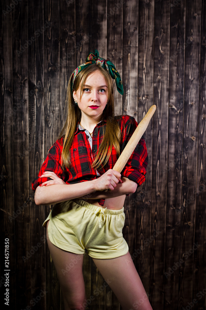 Obraz premium Young funny housewife in checkered shirt and yellow shorts pin up style with kitchen rolling pin on wooden background.