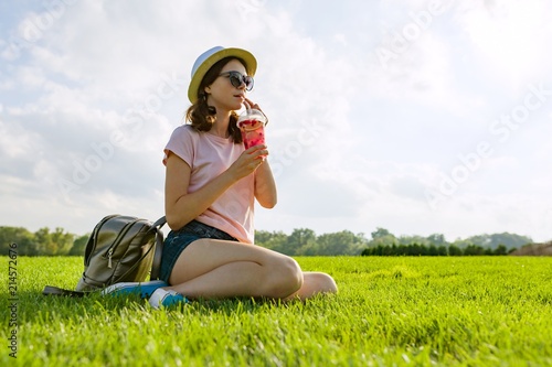 Young girl in sunglasses and hat drinks summer berry drink with ice sitting on green grass. Copy space. photo