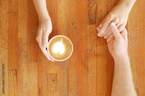 Young couple in love holding hands over red cup of coffee with heart shaped foam latte art on vintage grunged scratched table. Man & woman body language. Background, top view, close up, copy space.