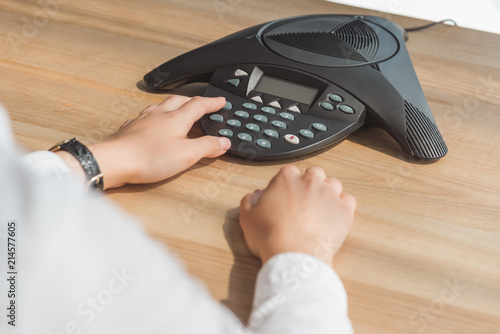 cropped shot of businesswoman pushing button of conference phone on table at office