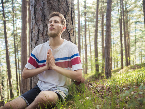 portrait of young bearded hipster doing yoga sitting under the tree in casual  concentration outdoor in forest park